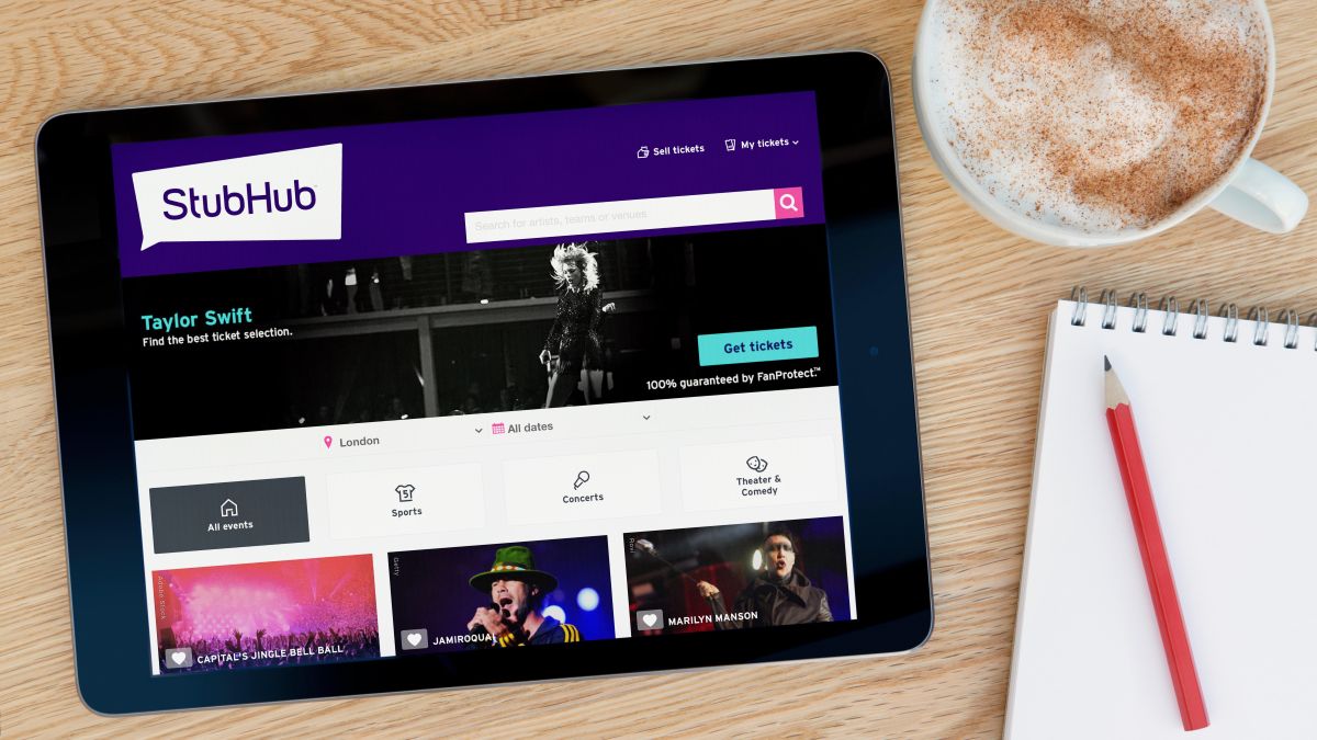 The Stubhub website features on an iPad tablet device which rests on a wooden table beside a notepad and pencil and a cup of coffee